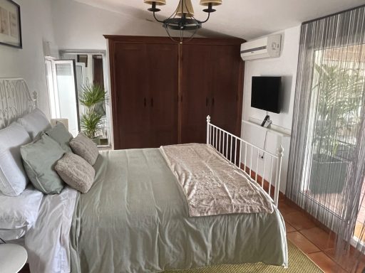Dormitorio con cama, armario de madera y televisión en la pared. Luz natural y decoración sencilla.