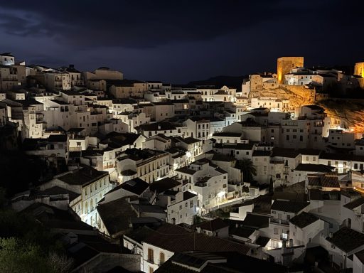 Vista nocturna de un pueblo blanco iluminado, con castillo en la colina.