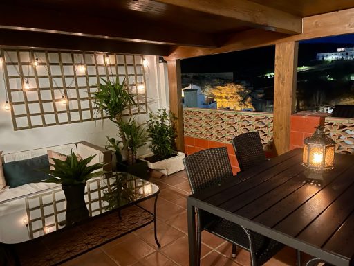 Terraza acogedora con plantas, mesa y sillas, iluminada con luces cálidas.