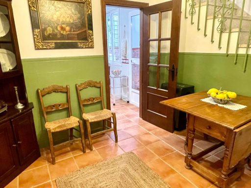 Entrante con paredes verdes, mesa de madera, sillas de mimbre y un cuadro en la pared.
