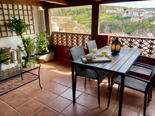 Terraza acogedora con mesa y sillas, bebida en mesa, vistas a un paisaje verde.