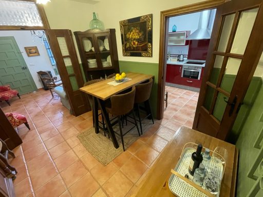 Cocina con mesa de madera, sillas y acceso a una habitación contigua. Decoración simple.