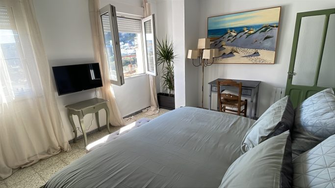 Habitación luminosa con cama, mesa de trabajo y televisor, decorada con un cuadro.