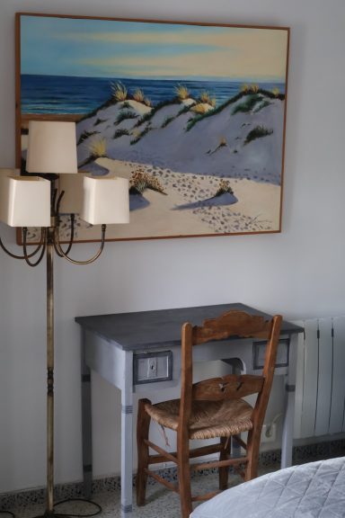 Escritorio gris con una silla de madera y una lámpara de pie, decorado con un cuadro de playa.